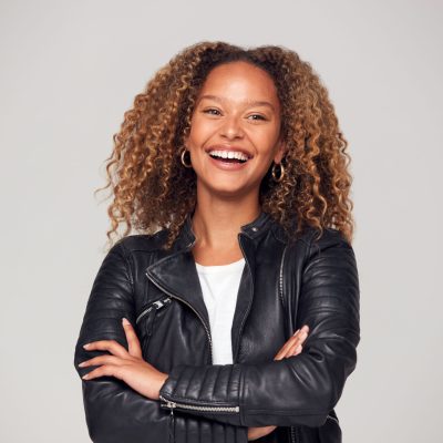 Waist Up Studio Shot Of Happy Young Woman With Folded Arms Wearing Leather Jacket Smiling At Camera